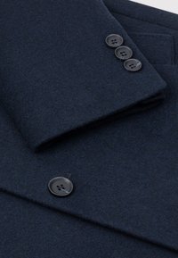 Granatowy wełniany płaszcz z założonym kołnierzem i trzema czarnymi guzikami na mankiecie. Tkanina ma miękkie, teksturowane wykończenie.