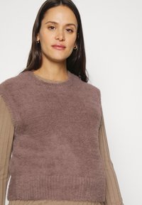 Sweater fofinho, sem mangas, na cor mauve suave, com uma textura macia e decote arredondado, sobre uma blusa de manga longa canelada.