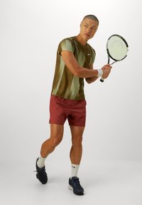Brun og grønn stripete treningstrøye, røde shorts, hvite sokker og mørke sko. Holder en tennisracket, i et halvveis sving.