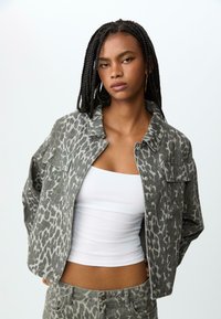 Jakna od trapera s leopard uzorkom u sivim nijansama, koja ima ovratnik, prednje džepove i širok kroj. Ukomponirana s bijelim uskim majicom bez rukava.