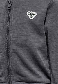 Grå zip-up jacka i texturerad tyg med en vit och grön broderad logotyp på vänster bröstområde.