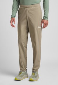 Pantaloni leggeri beige con cintura elastica, dotati di un piccolo logo bianco sulla coscia. Abbinati a scarpe da ginnastica grigie e gialle.
