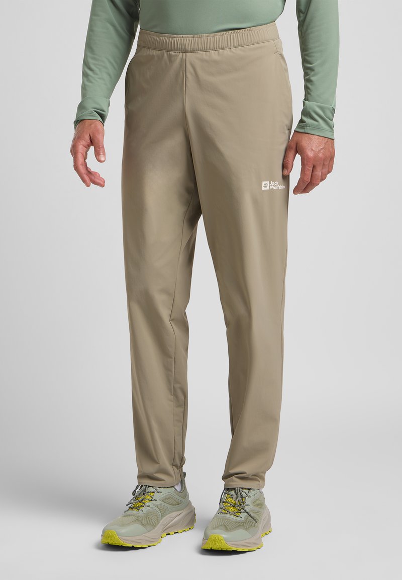 Beige leichte Hose mit elastischem Bund, mit einem kleinen weißen Logo an der Oberschenkelinnenseite. Kombiniert mit grauen und gelben Sportschuhen.