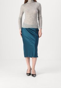 Femme portant un pull col roulé gris ajusté, une jupe midi en satin teal et des escarpins noirs à bout pointu, se tenant devant un fond blanc.