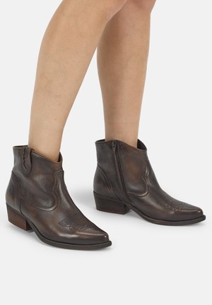 Beine mit dunkelbraunen Leder-Stiefeletten mit niedrigen Blockabsätzen und seitlichen Reißverschlüssen vor schlichtem hellem Hintergrund.