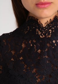 Svart spetsblus med blommönster, hög krage och delikat frans längs halsringningen; halvtransparenta tyget framhäver designens detaljer.