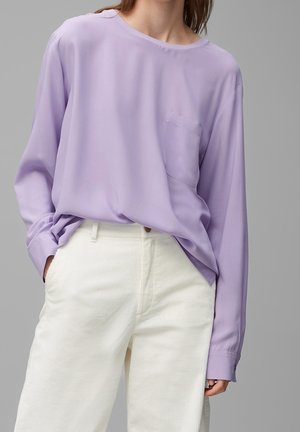 Personne portant une chemise ample lavande à manches longues avec une poche poitrine et un pantalon en velours côtelé blanc taille haute, debout avec une main dans la poche.