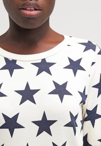 Sudadera blanca con patrón de estrellas en azul marino, presenta un cuello redondo y un acabado acanalado en el cuello. La tela parece suave y texturizada.