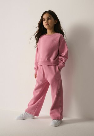 Ragazza giovane con lunghi capelli scuri in piedi, indossa una felpa rosa coordinata e pantaloni a gamba larga con scarpe da ginnastica bianche su sfondo semplice.