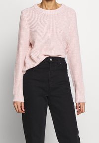 Hellrosa gestrickter Pullover mit Rundhalsausschnitt und langen Ärmeln, der ein strukturiertes Muster aufweist. Kombiniert mit hochgeschnittenen schwarzen Jeans.