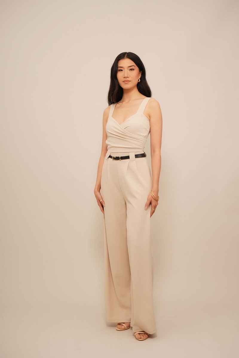 Combinaison beige avec un corps enroulé, jambes larges et une ceinture noire. Texture lisse, bretelles ajustables, légère plis sur le haut.