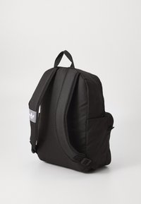 Sac à dos en tissu noir avec des bretelles rembourrées, une poche latérale et un logo sur un patch rectangulaire. Texture lisse et design minimaliste.