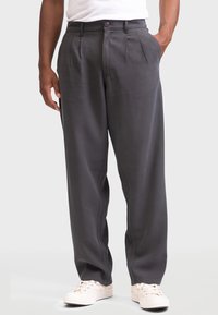 Hombre con pantalones plisados de color gris oscuro y zapatillas blancas, de pie con una mano en el bolsillo frente a un fondo liso.