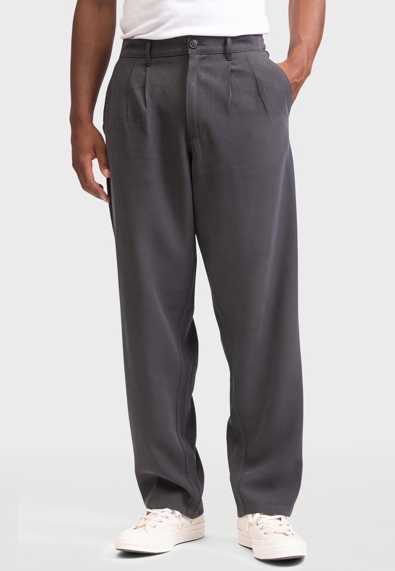 Hombre con pantalones plisados de color gris oscuro y zapatillas blancas, de pie con una mano en el bolsillo frente a un fondo liso.