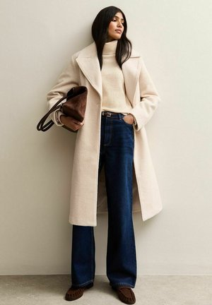 Largo abrigo crema, suéter de cuello alto, jeans de pierna ancha azul oscuro, bolso marrón. Tejido texturizado, líneas limpias y abrigo abierto que revela capas debajo.