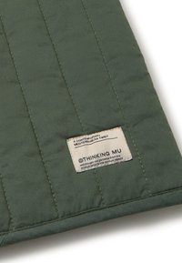 Gesteppt grüner Stoff mit vertikaler Naht; verfügt über ein beiges Etikett mit schwarzem Text und Logo von Thinking Mu. Weiche Textur, minimales Design.