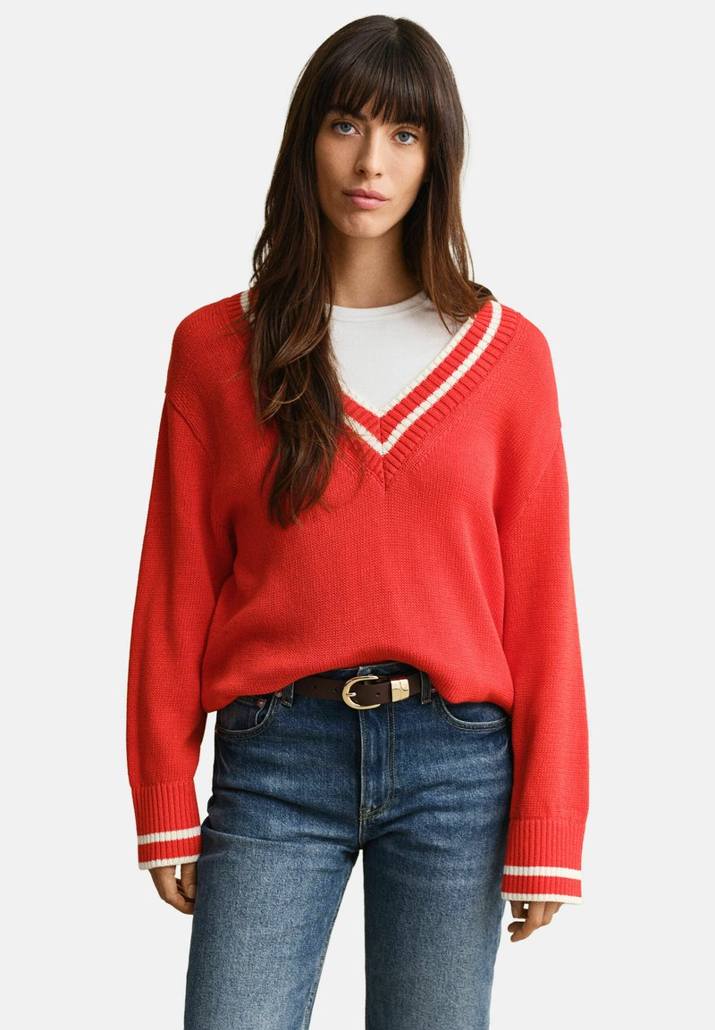 Femme aux longs cheveux noirs portant un pull rouge à col en V avec des rayures blanches, un sous-vêtement blanc, un jean bleu et une ceinture noire.