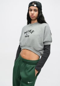 Γκρι cropped φούτερ με κεντημένο λογότυπο "Nike 1972", συνδυασμένο με σκούρο πράσινο παντελόνι φόρμας με λευκό λογότυπο Nike swoosh. Μαύρο καπέλο φορεμένο ανάποδα.