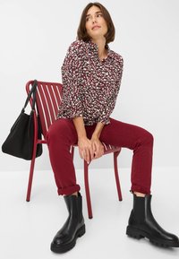 Camicia a fiori con tonalità di bordeaux, bianco e verde; abbinata a pantaloni bordeaux e stivali neri alla caviglia. Seduta su una sedia a strisce rosse.