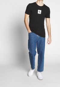 Camiseta negra de algodón con cuello redondo y logotipo blanco en el pecho, combinada con jeans azul denim de pierna recta y zapatillas blancas.