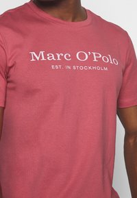 Różowa koszulka z krótkim rękawem, wykonana z miękkiej bawełny, z białym napisem: "Marc O'Polo" oraz "ZAŁ. W Sztokholmie." Klasyczny krój z okrągłym dekoltem.