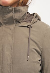 Olivově zelená bunda vyrobená z měkké tkaniny, se stahovací kapucí, šňůrkou s brzdičkou a hladkým povrchem. Přední zip je skrytý pod légou.