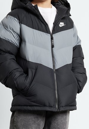Personne portant une doudoune Nike noire et grise avec capuche et fermeture éclair, les mains dans les poches, associée à un pantalon gris et une chemise blanche en dessous.