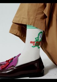 Chaussettes blanches côtelées avec des rayures vertes et un personnage vert avec le texte « Mr. Pickle », associées à un pantalon marron et des mocassins à pampilles rouge foncé.