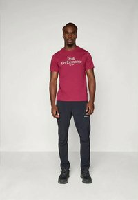 Maroon lyhythihainen t-paita, jossa on "Peak Performance" -logo. Tumma housut ja mustat kengät. Aseteltu neutraalia taustaa vasten.