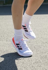 Białe buty sportowe z czarnymi i różowymi akcentami, wykonane z siateczkowego materiału, ze sznurówkami i gumową podeszwą. Noszone z białymi skarpetami do kostki.