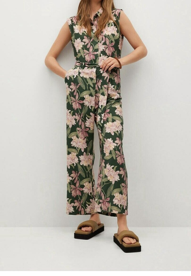 Femme portant une combinaison sans manches à motifs floraux verts, roses et beiges, debout les mains dans les poches et sandales compensées marron.