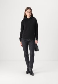 Haut à capuche noir avec un logo discret, associé à un jean gris foncé. Le mannequin porte des bottines noires élégantes et tient un petit sac à main noir.