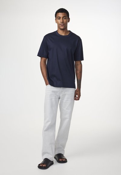 T-shirt in cotone blu navy con vestibilità rilassata, maniche corte e scollo rotondo. Indossato con jeans dritti grigi chiari e sandali neri.