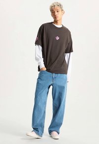 Dark brown t-shirt with a pink floral patch, layered over a white long-sleeve top. Paired with light blue baggy jeans and checkered slip-on shoes.