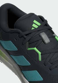 Baskets de sport noires avec une tige en mesh texturé, accents verts et bleus, col rembourré et lacets gris. Présente un motif de logo de trois bandes.