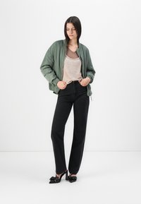 Grønn bomberjakke, beige topp, høyt liv svarte jeans og svarte hælesko med sløyfedetaljer, stående mot en enkel hvit bakgrunn.