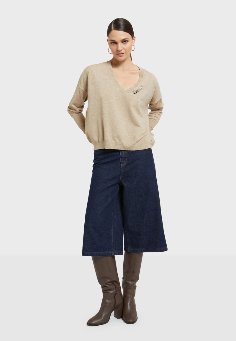 Bež pleteni pulover s V-izrezom i slobodnim krojem, u kombinaciji s tamnoplave široke traperice do koljena i smeškim čizmama s petom do koljena.