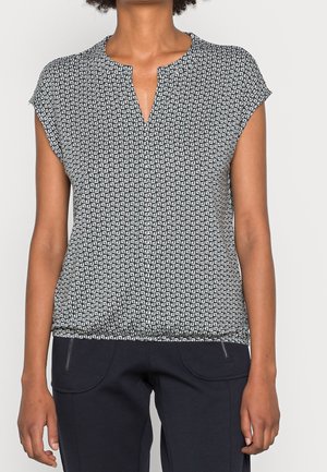 Femme portant un haut à manches courtes à motif géométrique noir et blanc avec un petit col en V et un pantalon noir avec des poches zippées.