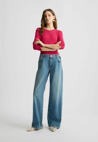 Rotes Langarmoberteil kombiniert mit weiten, hellblauen Jeans. Auffällige Merkmale sind die taillierte Taille und die strukturierte Silhouette. Peeptoe-Schuhe.