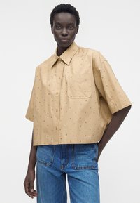 Persona con una camisa beige suelta de manga corta con pequeños detalles y un bolsillo en el pecho, combinada con vaqueros azules, de pie con una mano en el bolsillo.