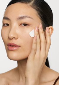 Fuktighetskrem påføres kinnet; produktet er hvitt og kremete. Modellen har rett, mørkt hår og lys hud.