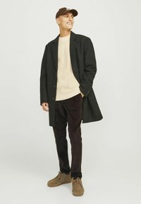 Un manteau en laine noir porté par-dessus un pull en laine beige, un pantalon en velours côtelé marron foncé et des chaussures marron. La casquette est marron foncé avec un logo.