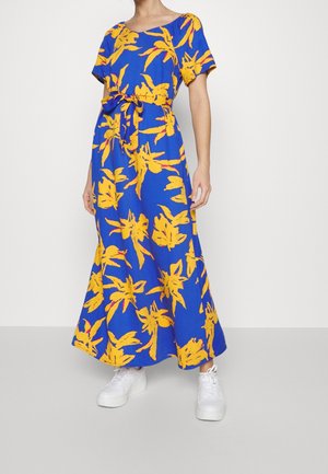 Niebieska sukienka maxi w żółty kwiatowy wzór z paskiem w talii, noszona z białymi sneakersami na stojącej osobie na jednolitym tle.