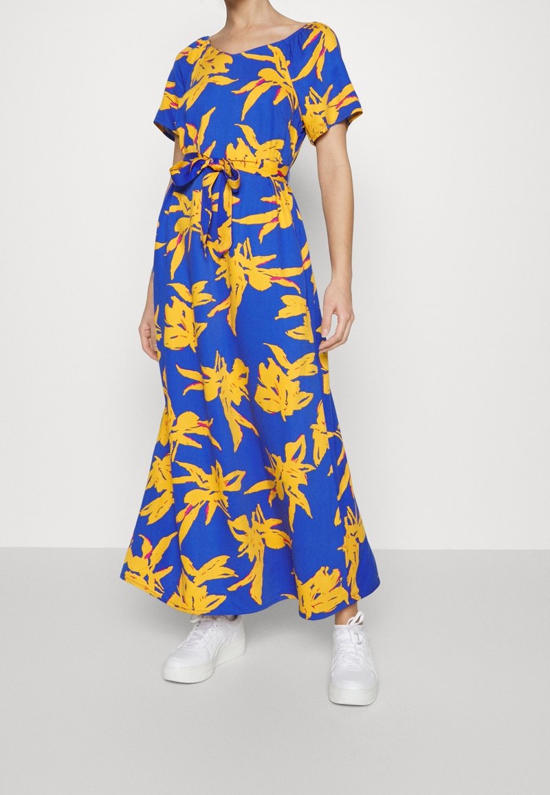 Robe maxi bleue avec imprimé floral jaune et ceinture à la taille, portée avec des baskets blanches par une personne debout sur un fond uni.