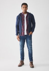 Marineblå zip-up hættetrøje over en rød og blå ternet skjorte, hvid t-shirt, blå jeans og brune læderstøvler, stående mod en ensfarvet baggrund.