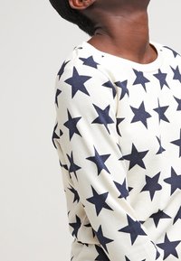Sudadera blanca con estampado de estrellas azul marino, cuello redondo y mangas largas. Tejido suave con un corte relajado y detalles de costura sutiles.
