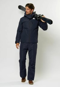 Mann in navyblauer Skijacke und -hose, der Skier über der Schulter trägt, mit braunen Stiefeln, vor einem hellen, einfarbigen Hintergrund stehend.
