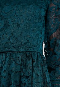 Tauchblaues Spitzenkleid mit aufwendigen Blumenmustern, langen Ärmeln und einem gerafften Rock. Das Material hat ein strukturiertes, schichtartiges Aussehen.