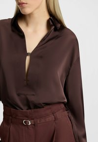 Camicia in satin marrone con scollatura a V profonda, un sottile ritaglio frontale e un dettaglio metallico decorativo al colletto. Abbinata a pantaloni coordinati.