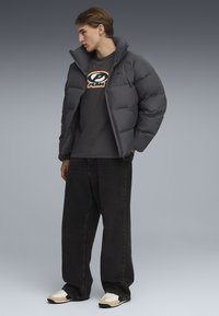 Grijze pufferjack met een hoge kraag, gedragen over een donker T-shirt met een logo. Gecombineerd met losse zwarte jeans en sneakers met beige accenten.